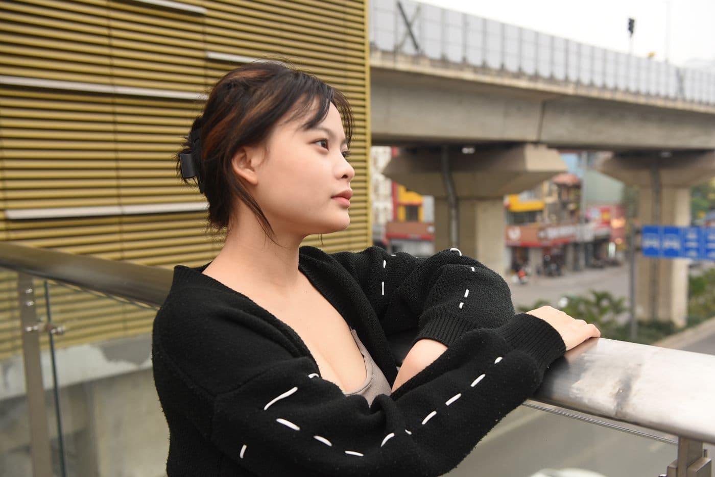 ผู้หญิงสวมคาร์ดิแกนไหมพรมสีดำ ยืนอยู่บนทางเดินลอยฟ้าในเมือง